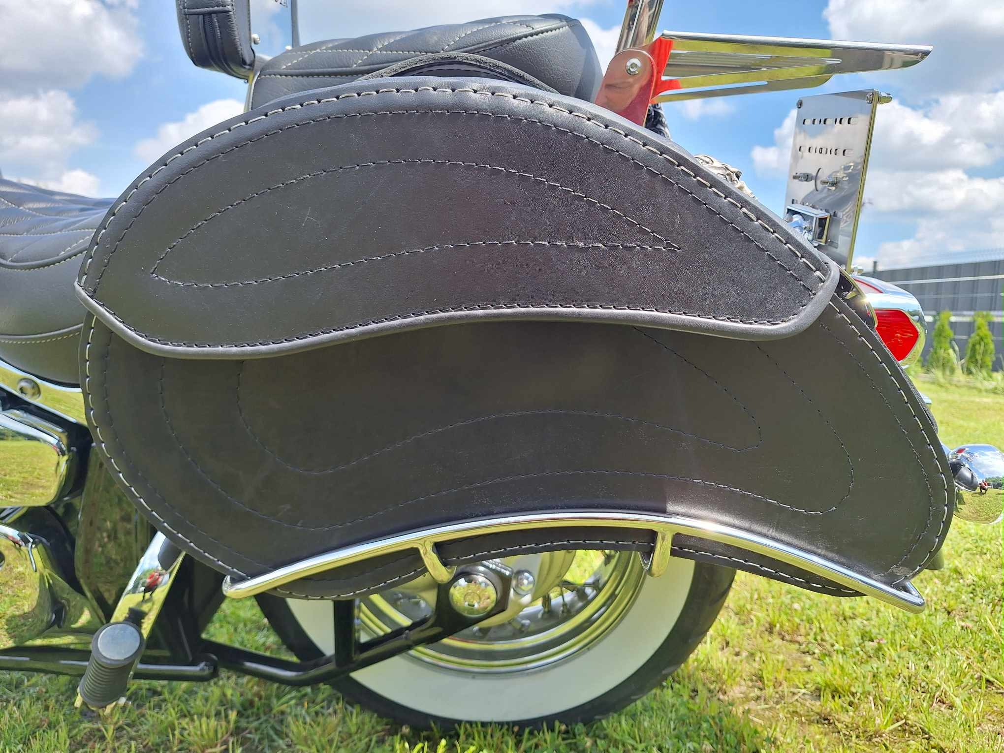 Czarna, skórzana sakwa motocyklowa z białymi przeszyciami, zamontowana na chromowanym stelażu, widoczna na tle zielonej trawy i błękitnego nieba w Jedlińsku.