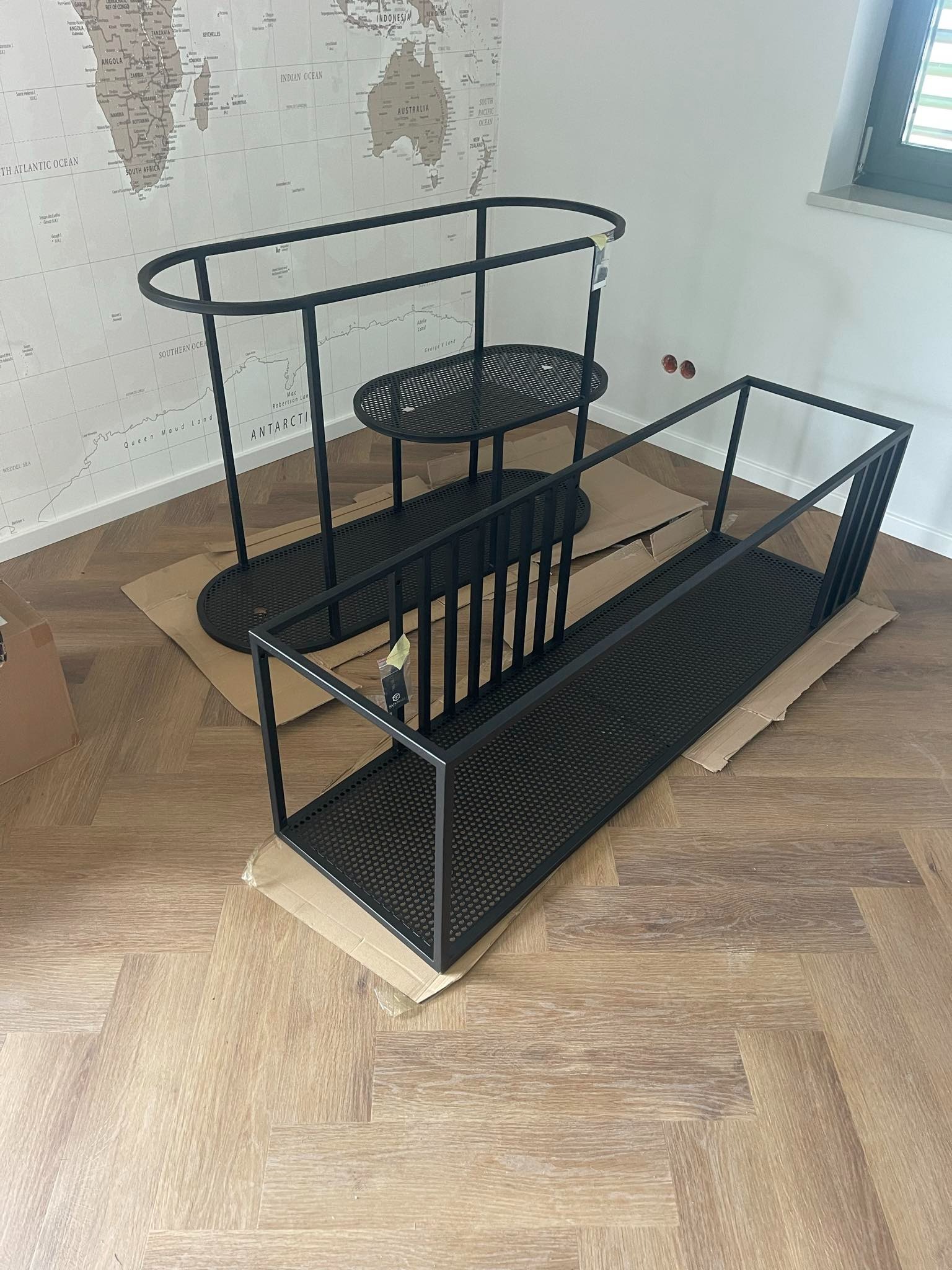 Dwa czarne, metalowe elementy konstrukcyjne, możliwe elementy balustrady lub ogrodzenia, leżące na kartonie na drewnianej podłodze, z widoczną mapą świata na ścianie w tle.