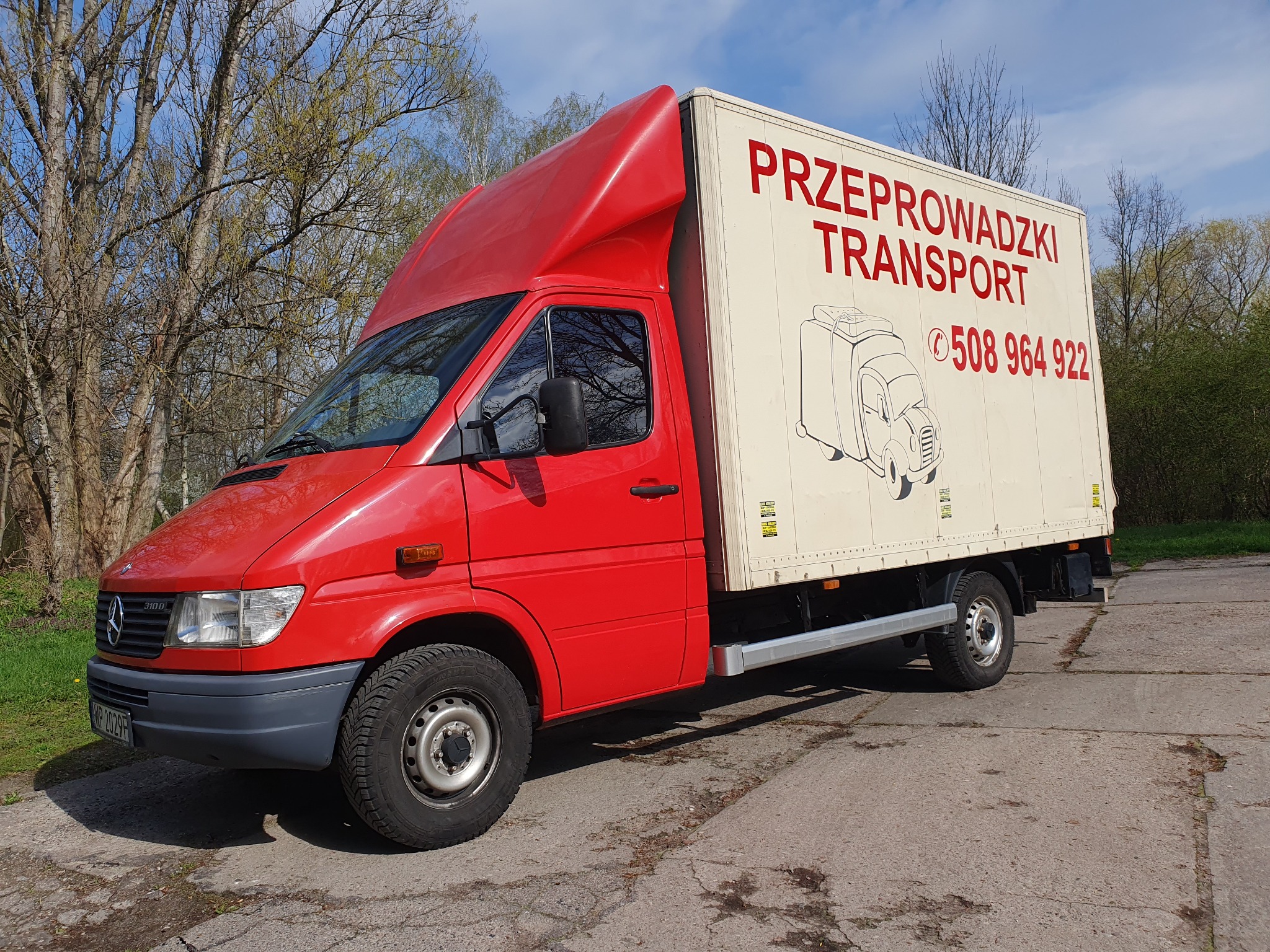 Czerwony samochód dostawczy z nadrukiem 'Przeprowadzki Transport' i numerem telefonu, zaparkowany na betonowym placu z drzewami w tle.