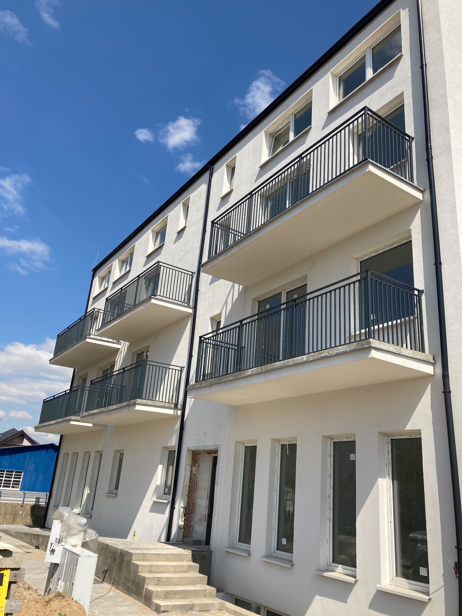 Nowo wybudowany budynek mieszkalny z balkonami zabezpieczonymi czarnymi, metalowymi balustradami, widoczne schody wejściowe i elementy wykończeniowe.