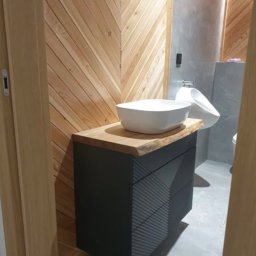 Meble inny wymiar - Nowoczesna łazienka z drewnianą ścianą w jodełkę, czarną szafką umywalkową z drewnianym blatem i białą umywalką nablatową. Widoczny fragment toalety.