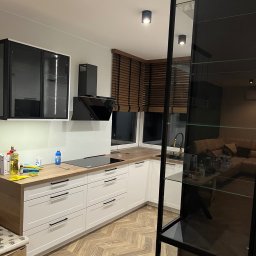 Meble inny wymiar - Nowoczesna kuchnia w bieli i drewnie: białe fronty szafek z czarnymi uchwytami, drewniany blat, czarna płyta indukcyjna i okap. Parkiet w jodełkę na podłodze.