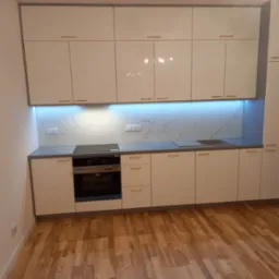 Biała, nowoczesna zabudowa kuchenna z połyskującymi frontami, złotymi uchwytami, blatem w odcieniach szarości i marmurowym backsplashiem podświetlonym niebieskim światłem LED, widoczna płyta...