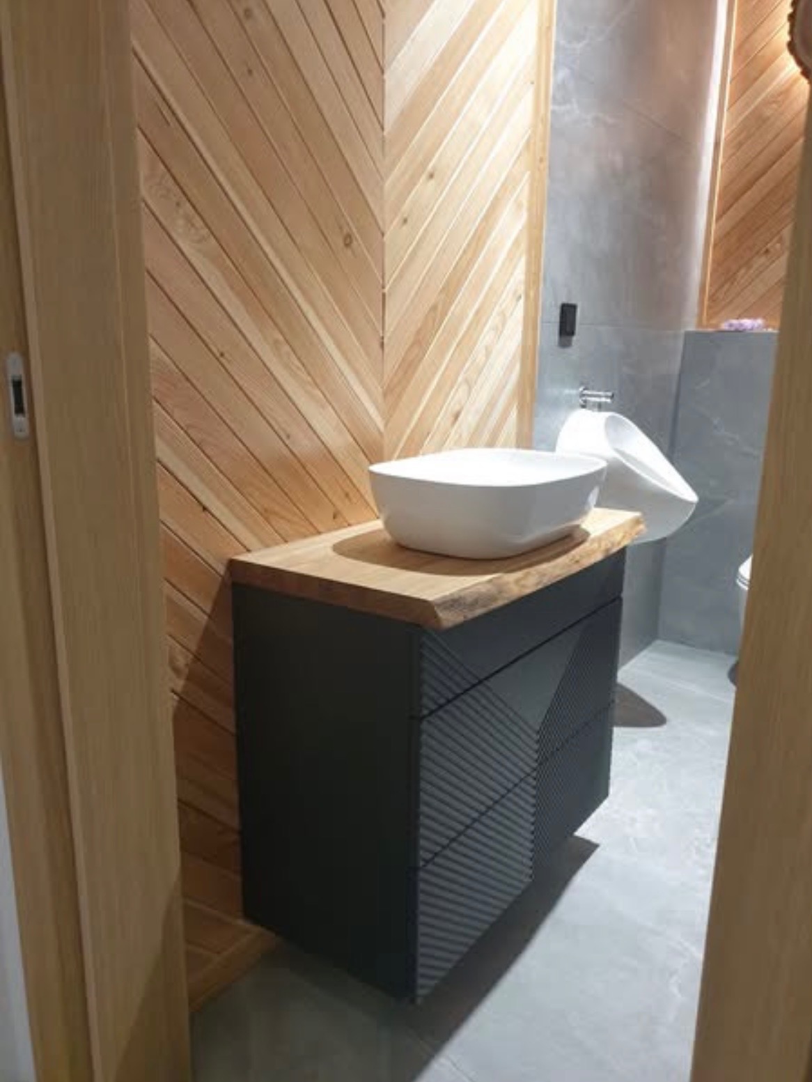 Nowoczesna łazienka z drewnianą ścianą w jodełkę, czarną szafką umywalkową z drewnianym blatem i białą umywalką nablatową. Widoczny fragment toalety.