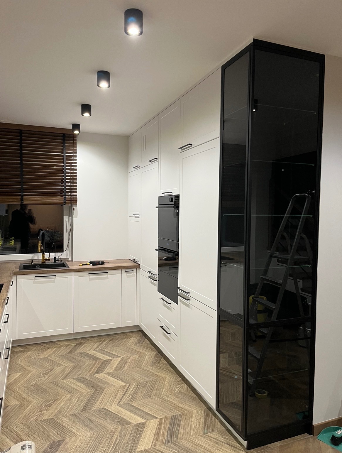 Nowoczesna kuchnia na wymiar z białymi frontami, czarnymi uchwytami i wysoką, przeszkloną szafką. Parkiet w jodełkę na podłodze. Widoczne oświetlenie sufitowe.