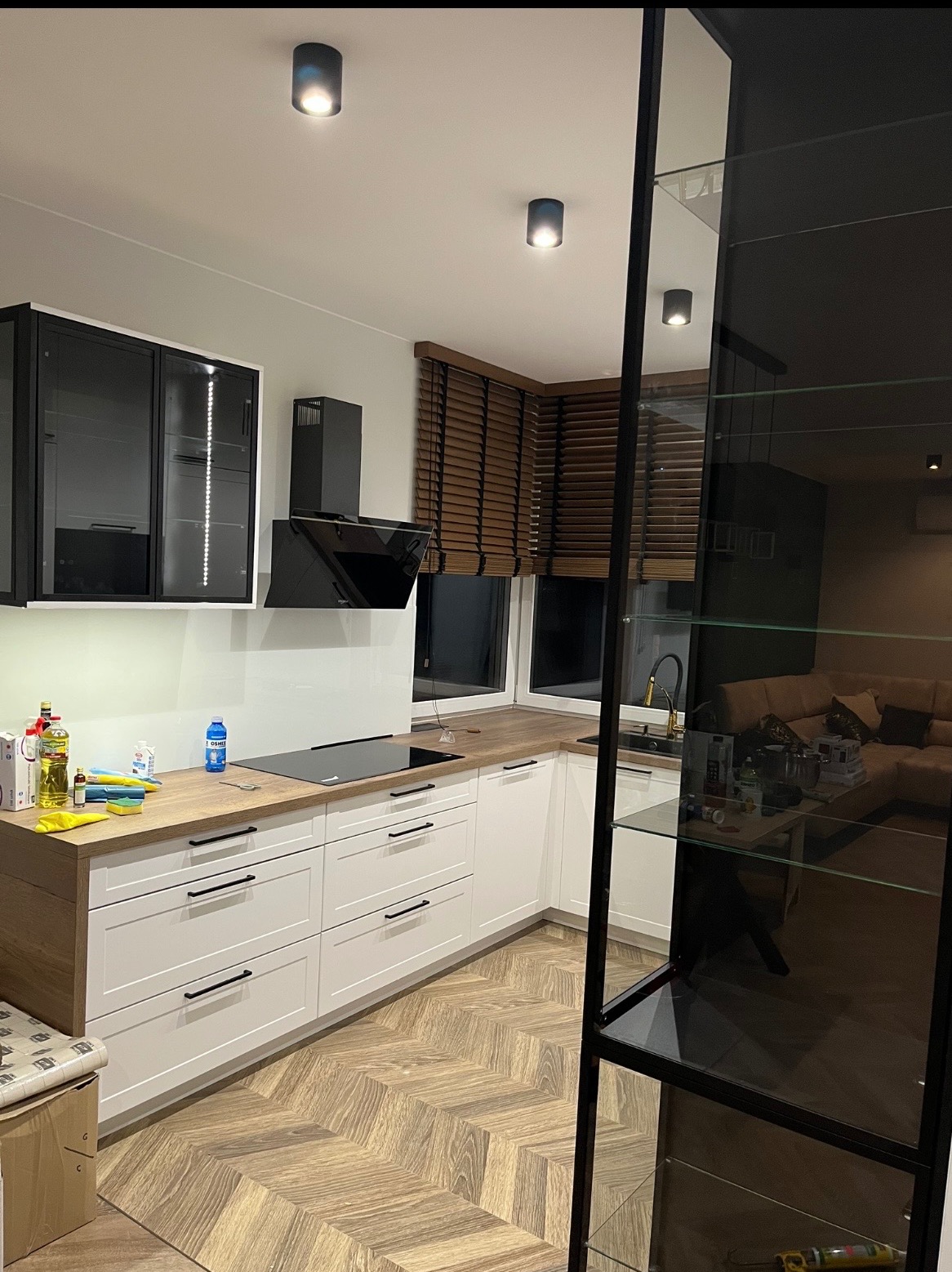 Nowoczesna kuchnia w bieli i drewnie: białe fronty szafek z czarnymi uchwytami, drewniany blat, czarna płyta indukcyjna i okap. Parkiet w jodełkę na podłodze.