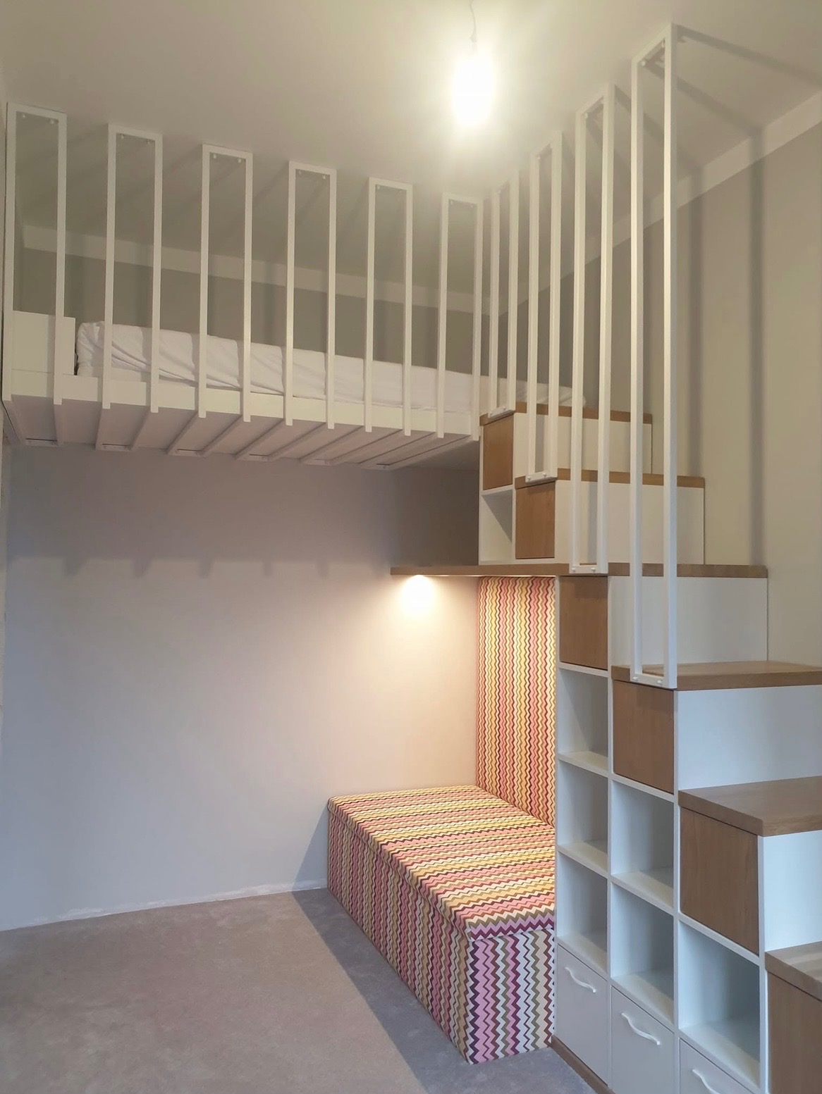 Pokój z antresolą: białe, minimalistyczne meble na wymiar, schody z szafkami i tapicerowane siedzisko w zygzaki. Oświetlenie punktowe podkreśla detale.