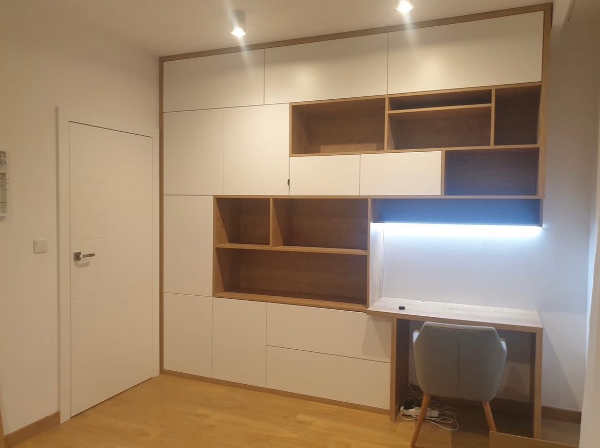 Biało-drewniana szafa wnękowa z biurkiem i oświetleniem LED, nowoczesny design, widok na całą ścianę, szara krzesło biurowe, jasna podłoga.