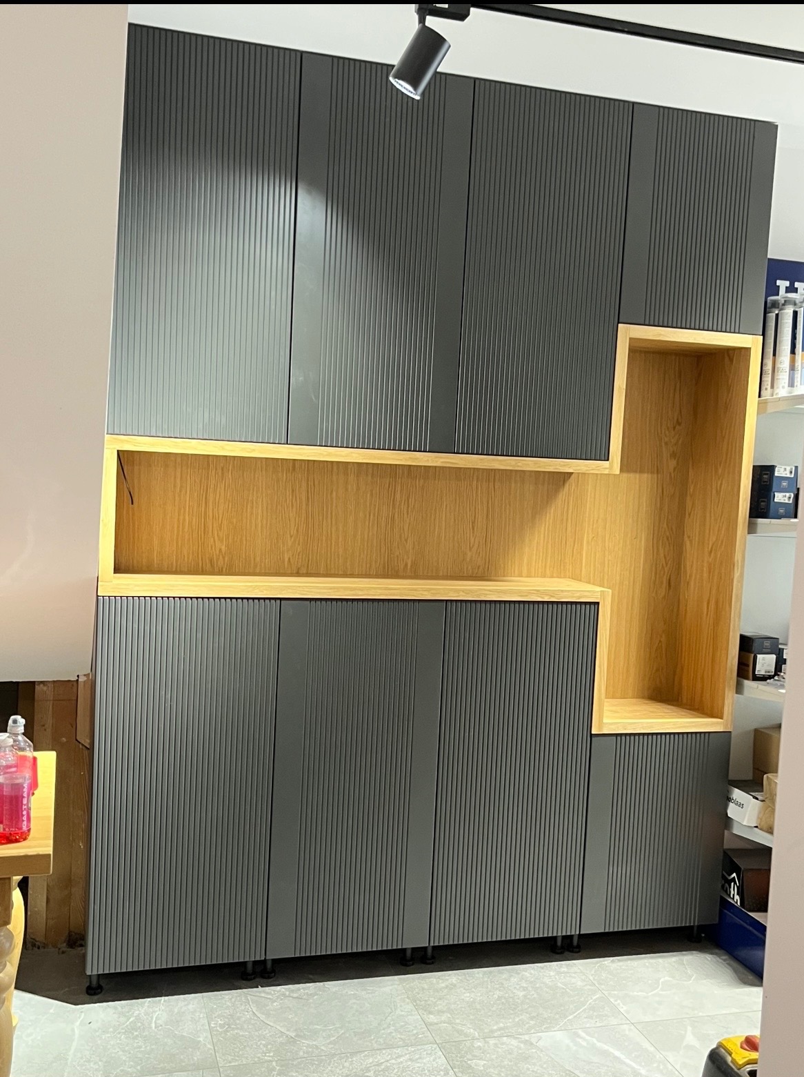 Szara szafa wnękowa z ryflowanymi frontami i drewnianymi wstawkami, oświetlona punktowo, na tle jasnej ściany i podłogi z płytek. Minimalistyczny design.