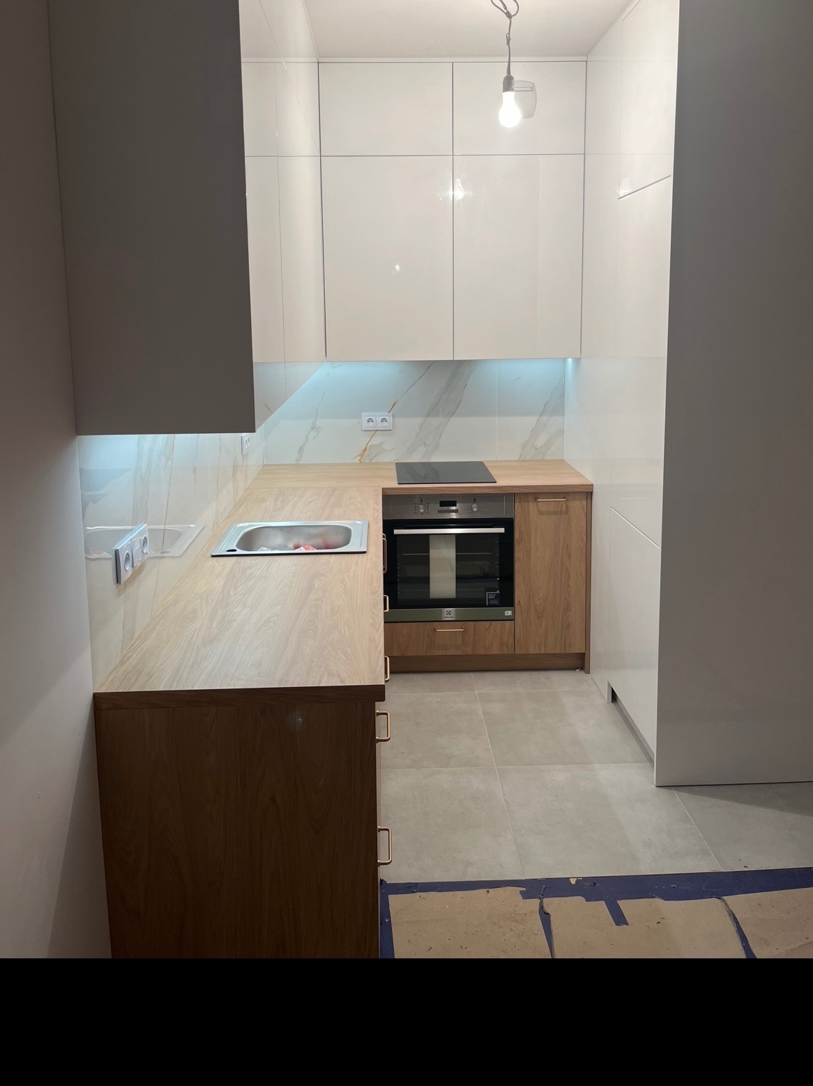 Jasna kuchnia z drewnianymi akcentami: zabudowa z białymi szafkami i drewnianym blatem, oświetlenie LED pod szafkami, płyta indukcyjna i piekarnik w zabudowie.