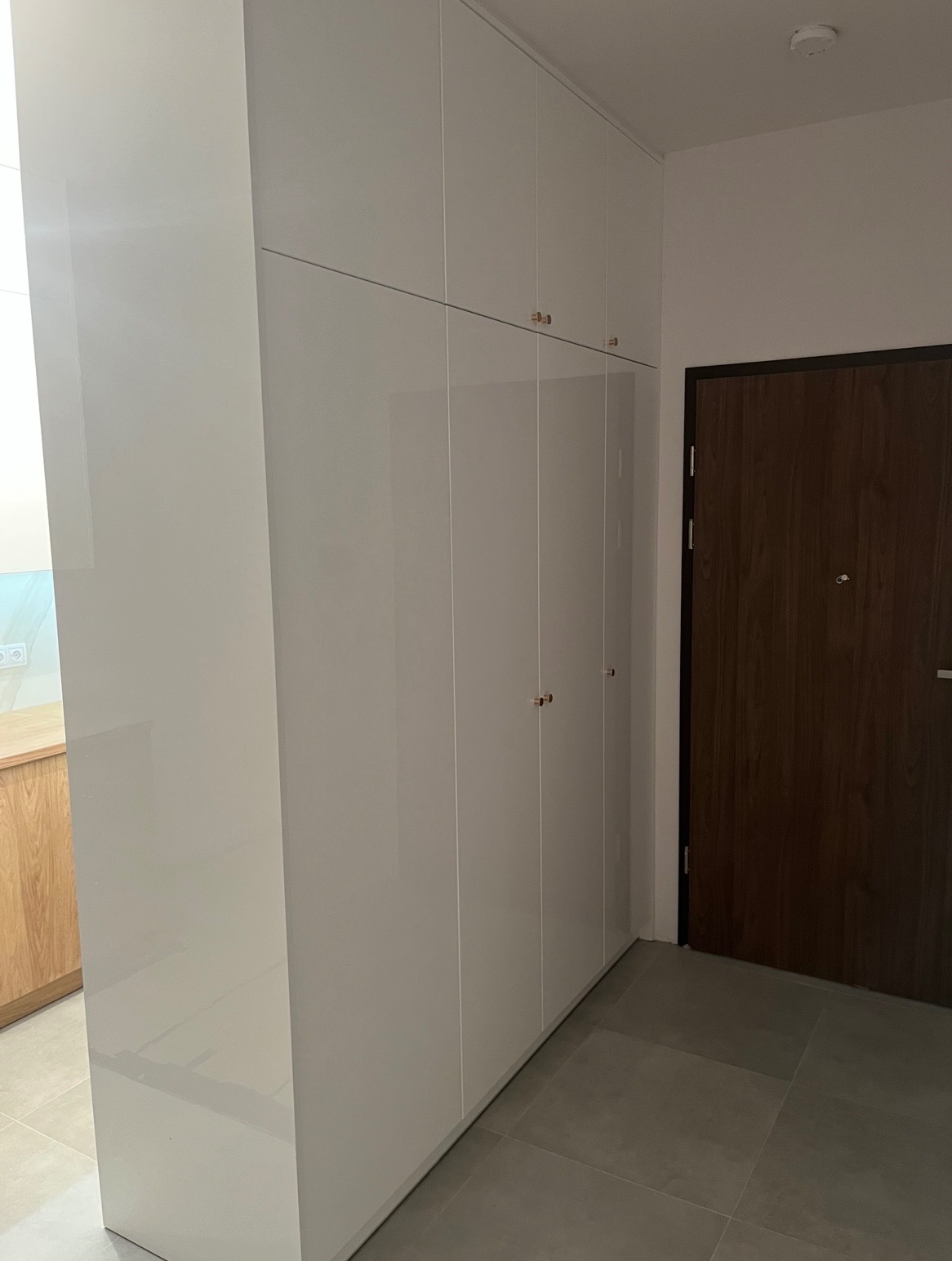 Biała, wysoka szafa wnękowa z połyskiem, z delikatnymi złotymi uchwytami, dopasowana do wnętrza, widoczna obok drewnianych drzwi i fragmentu kuchni.