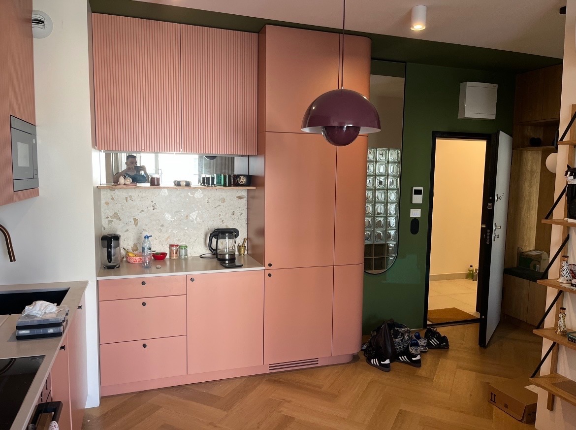 Różowa zabudowa kuchenna z ryflowanymi szafkami górnymi i blatem z lastryko. W tle widoczne lustro i wejście do innego pomieszczenia z luksferami.