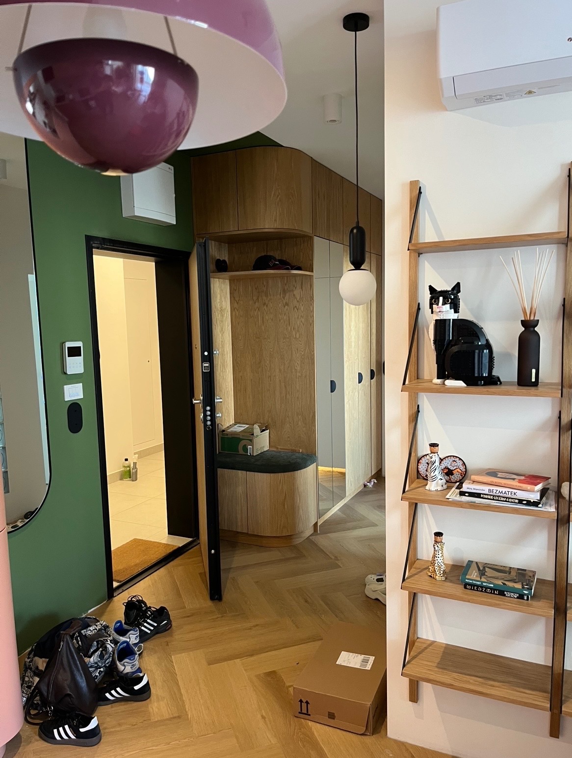 Nowoczesny przedpokój z zabudową z jasnego drewna, otwartymi szafkami i siedziskiem. Na podłodze parkiet w jodełkę. Dekoracyjna półka z książkami i ozdobami.