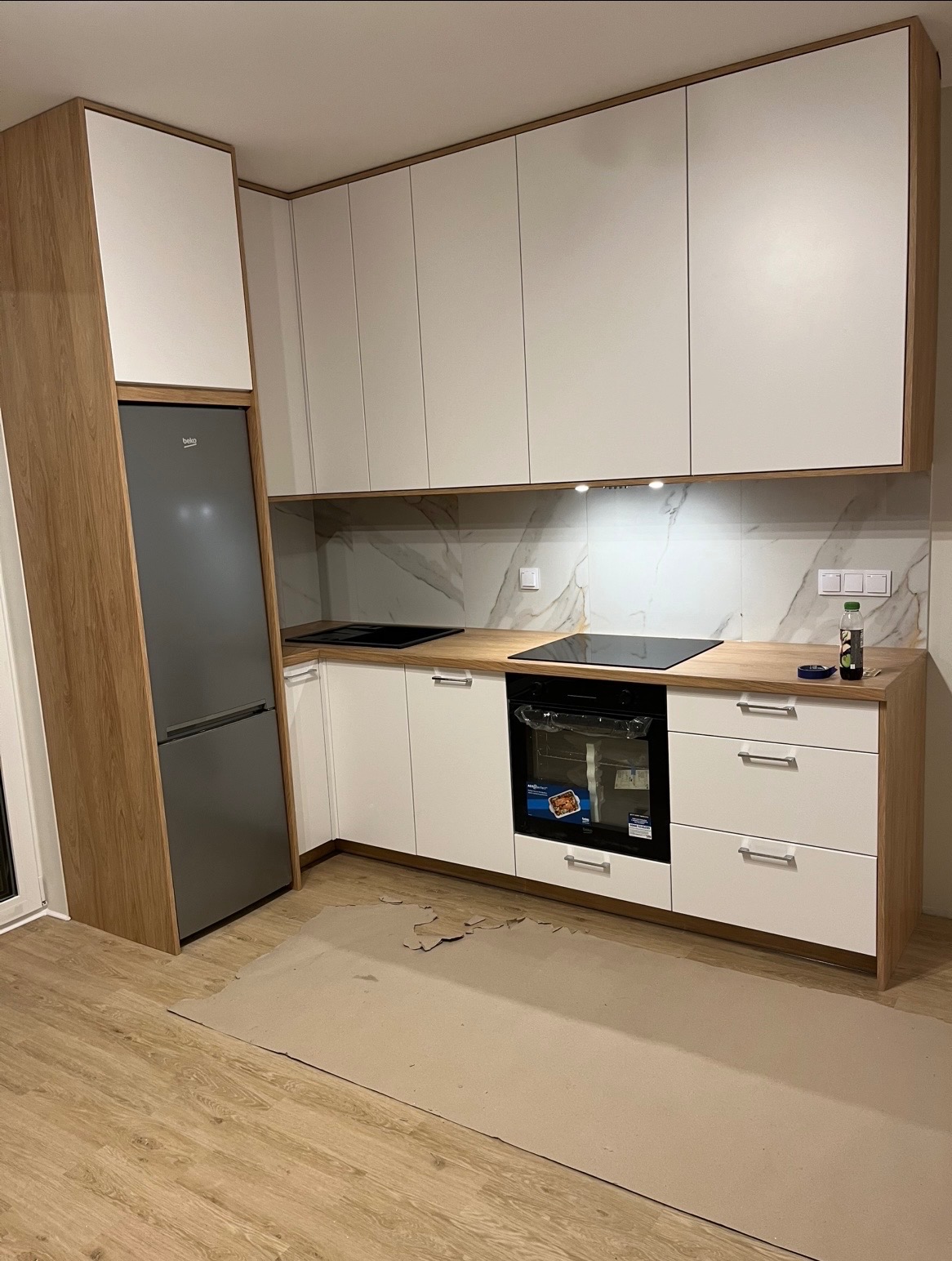 Nowoczesna kuchnia z białymi frontami i drewnianymi akcentami. Zabudowa z lodówką, piekarnikiem i płytą indukcyjną. Marmurowy backsplash i drewniany blat. Podłoga wyłożona panelami.