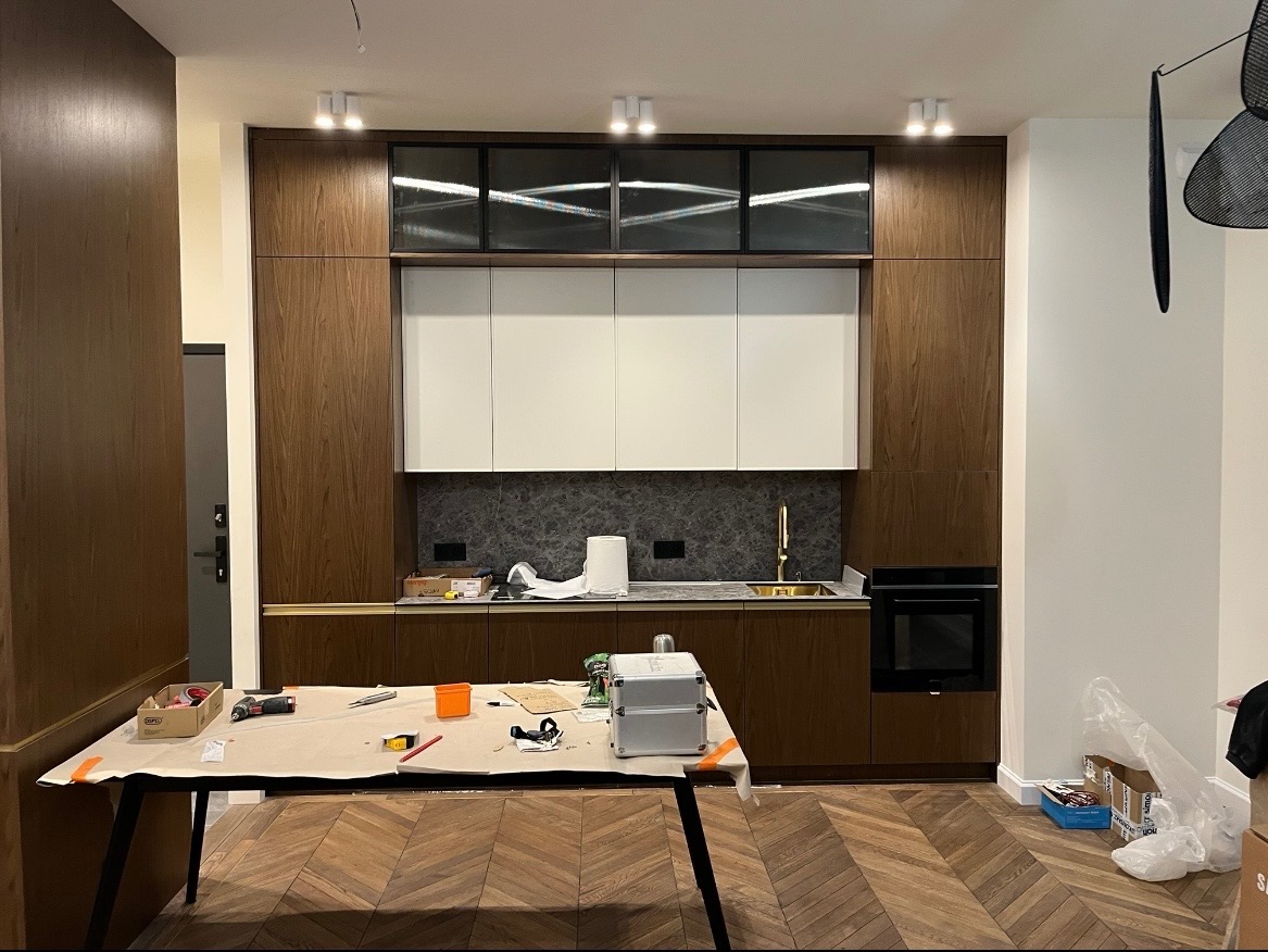 Nowoczesna kuchnia z drewnianymi frontami i białymi szafkami wiszącymi. Na pierwszym planie stół roboczy z narzędziami, parkiet w jodełkę. Oświetlenie punktowe.