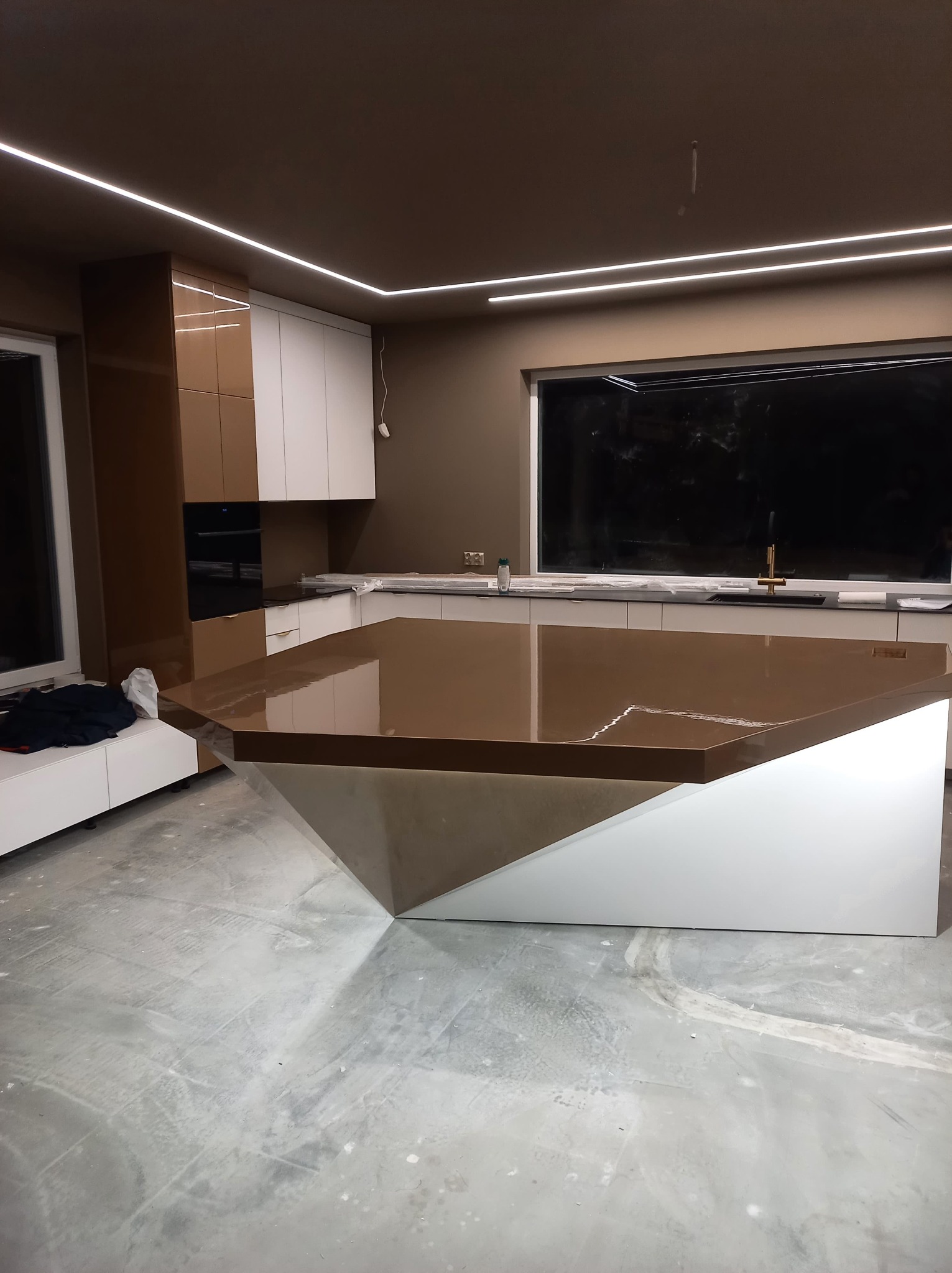 Wnętrze kuchni w trakcie montażu z wyspą o nietypowym kształcie, brązowy blat z połyskiem, białe szafki i oświetlenie LED podszafkowe, widoczne ślady po remoncie na podłodze.