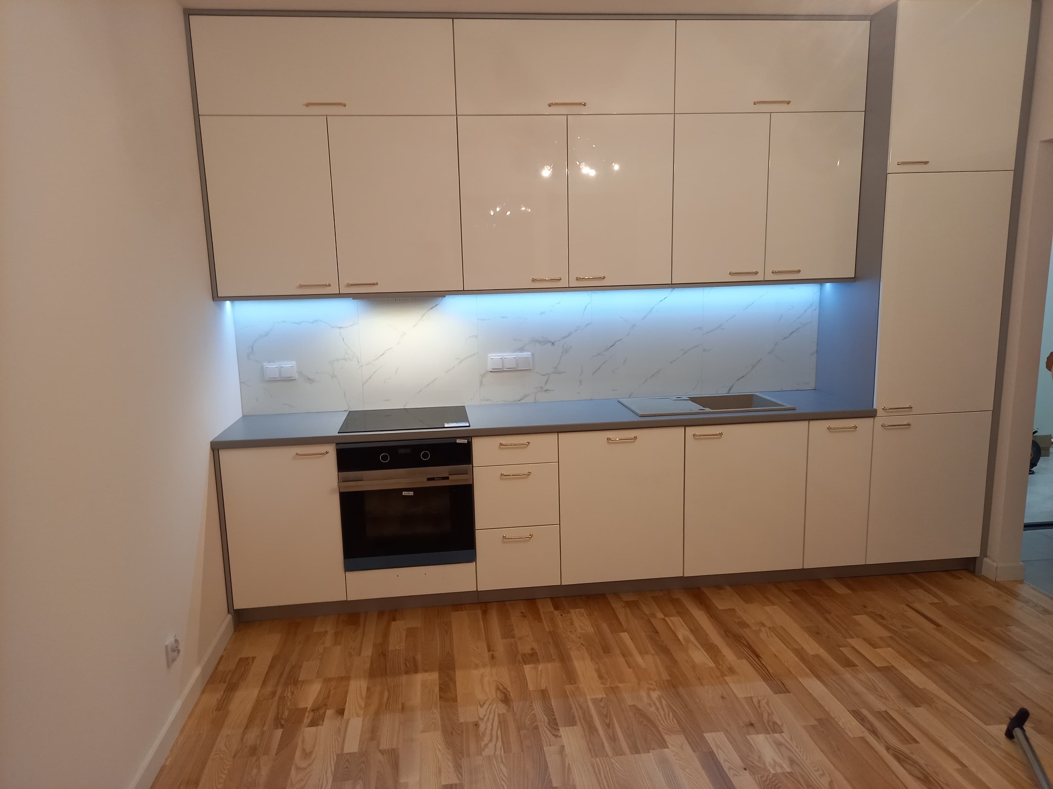 Biała, nowoczesna zabudowa kuchenna z połyskującymi frontami, złotymi uchwytami, blatem w odcieniach szarości i marmurowym backsplashiem podświetlonym niebieskim światłem LED, widoczna płyta...