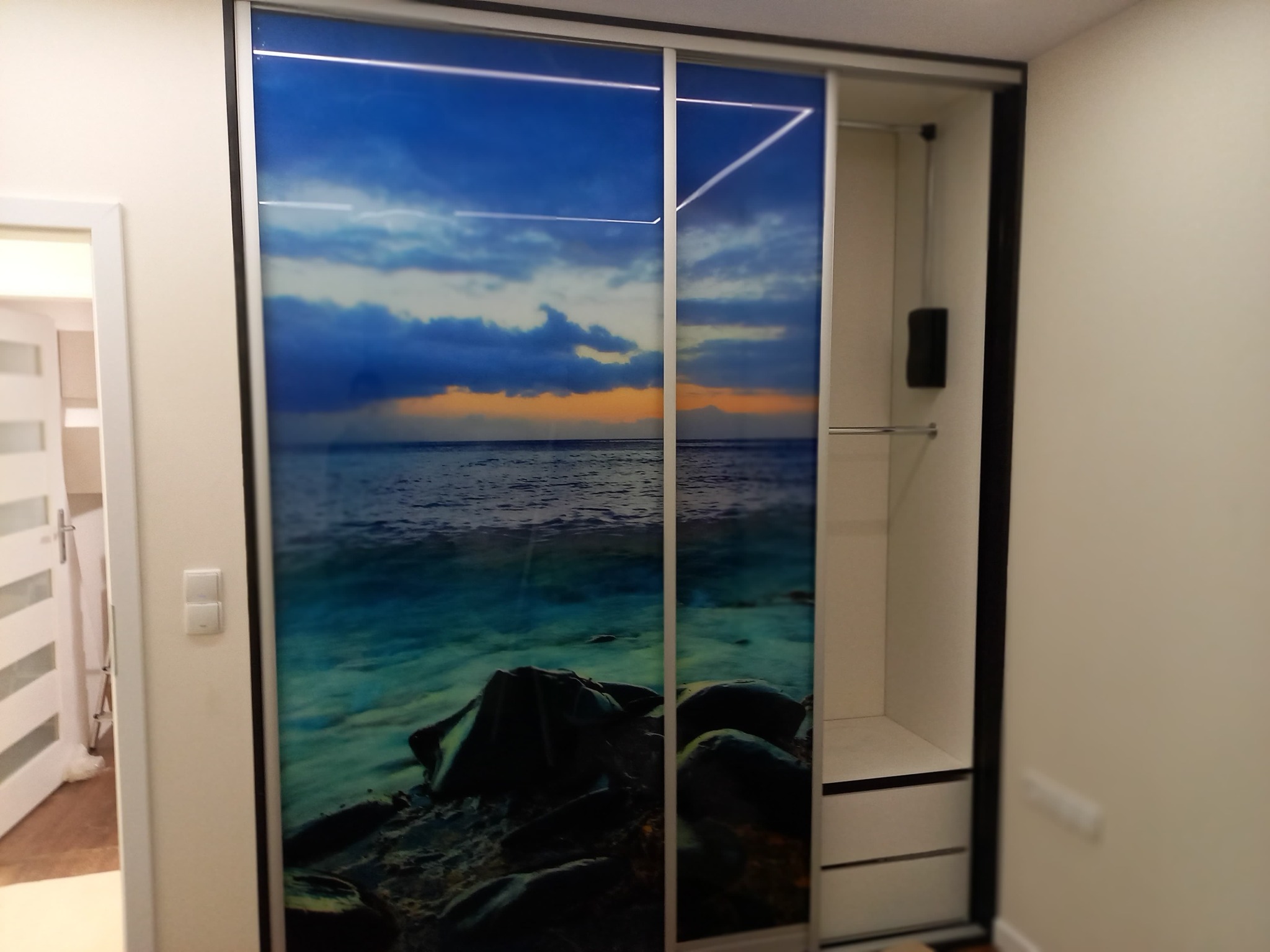Szafa przesuwna z fototapetą przedstawiającą morski krajobraz z kamienistym brzegiem i zachodem słońca, widoczna wnęka z półkami i drążkiem na ubrania.