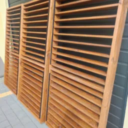 Trzy drewniane, brązowe konstrukcje z poziomymi lamelami, oparte o szarą fasadę z blachy trapezowej i ułożone na szarej kostce brukowej.