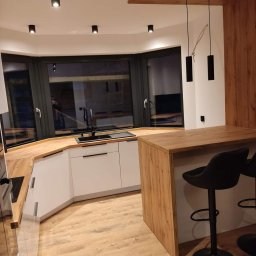 Drew Home Anna Przytuła - Nowoczesna kuchnia z białymi szafkami, drewnianym blatem i barkiem, oświetlona wiszącymi lampami i punktowymi reflektorami, z widocznym oknem wykuszowym.