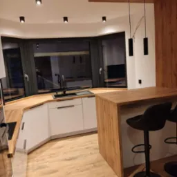 Nowoczesna kuchnia z białymi szafkami, drewnianym blatem i barkiem, oświetlona wiszącymi lampami i punktowymi reflektorami, z widocznym oknem wykuszowym.