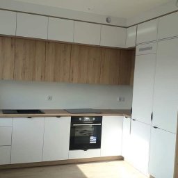 Drew Home Anna Przytuła - Nowoczesna kuchnia w kształcie litery L z białymi szafkami dolnymi i górnymi, z drewnianymi frontami szafek wiszących, czarnym piekarnikiem i płytą indukcyjną, oraz drewnianym blatem.