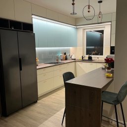 Drew Home Anna Przytuła - Nowoczesna kuchnia z jasnymi szafkami, turkusowym panelem ściennym, drewnianym blatem i barkiem oraz trzema designerskimi lampami wiszącymi.