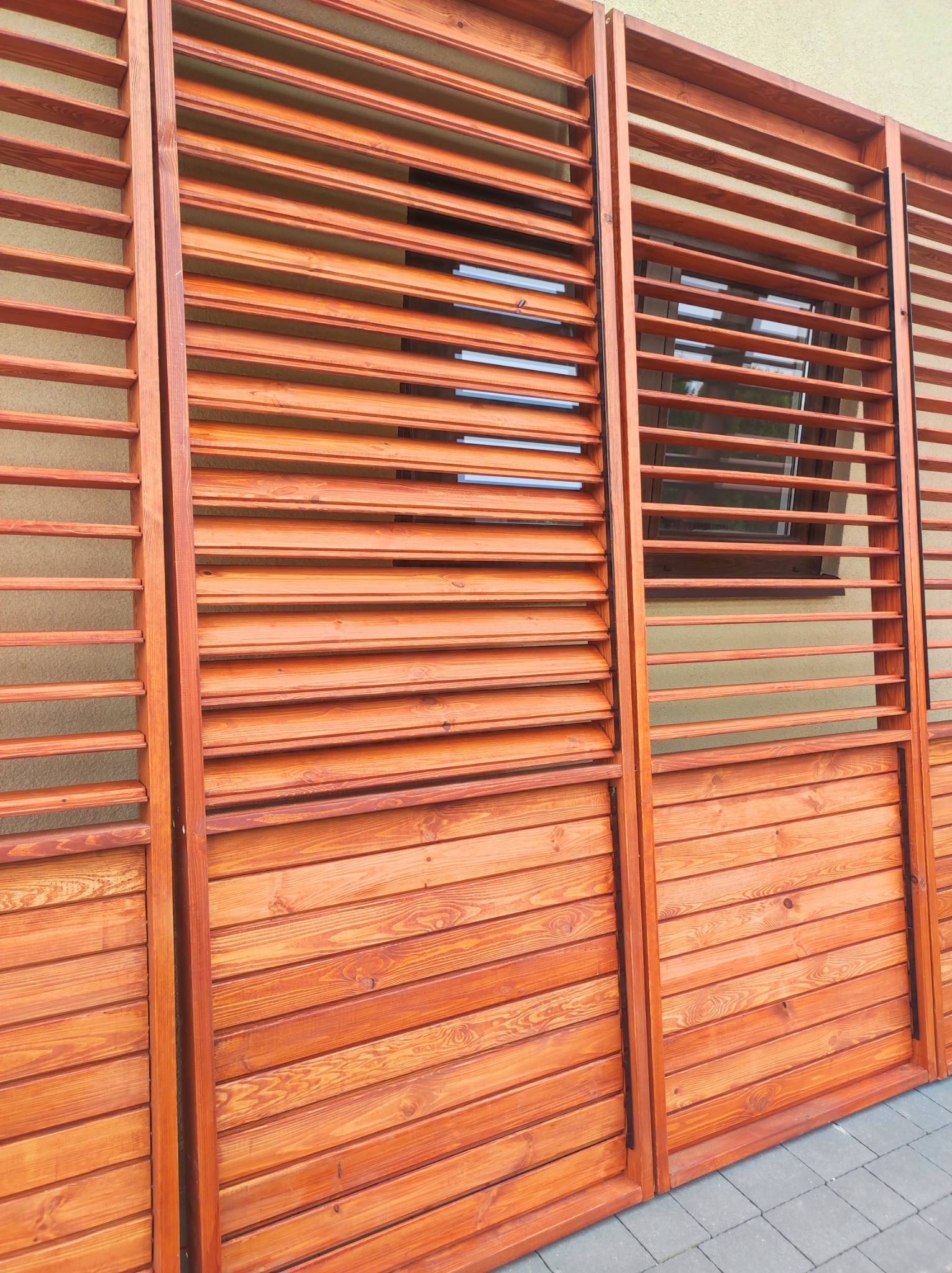 Trzy drewniane panele żaluzjowe w odcieniach brązu, ustawione obok siebie, częściowo zasłaniające okno w budynku o jasnej elewacji.