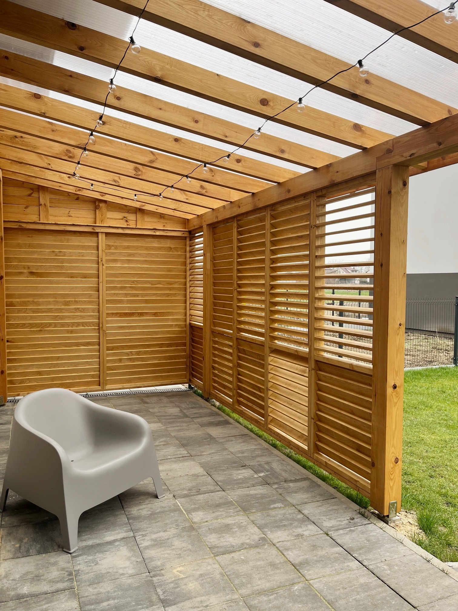 Drewniana konstrukcja z zadaszeniem z poliwęglanu, oświetlona girlandą żarówkową, z drewnianymi panelami żaluzjowymi na ścianach bocznych i szarą kostką brukową na podłodze, z szarym krzesłem...