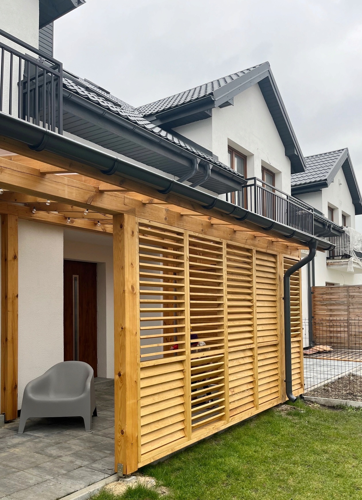 Drewniana konstrukcja z lamelami przy nowoczesnym domu jednorodzinnym, szara plastikowa krzesło na kostce brukowej, oświetlenie w postaci sznura żarówek.