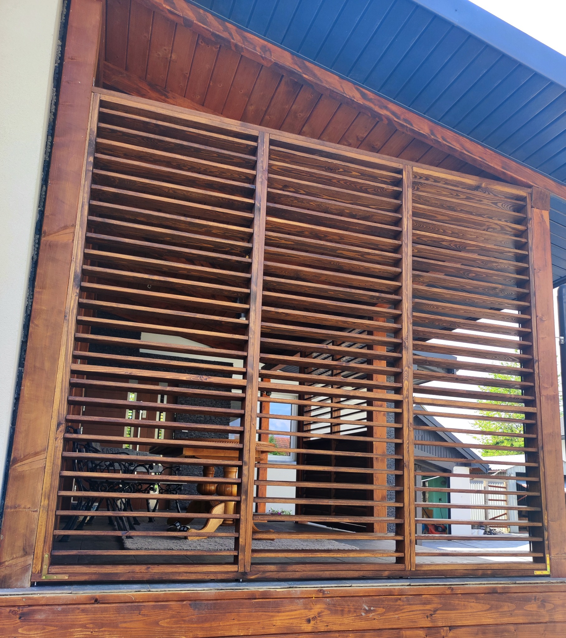 Drewniana konstrukcja z poziomych listew, tworząca ażurową ścianę, z widocznym wnętrzem z meblami ogrodowymi i elementami otoczenia na zewnątrz.