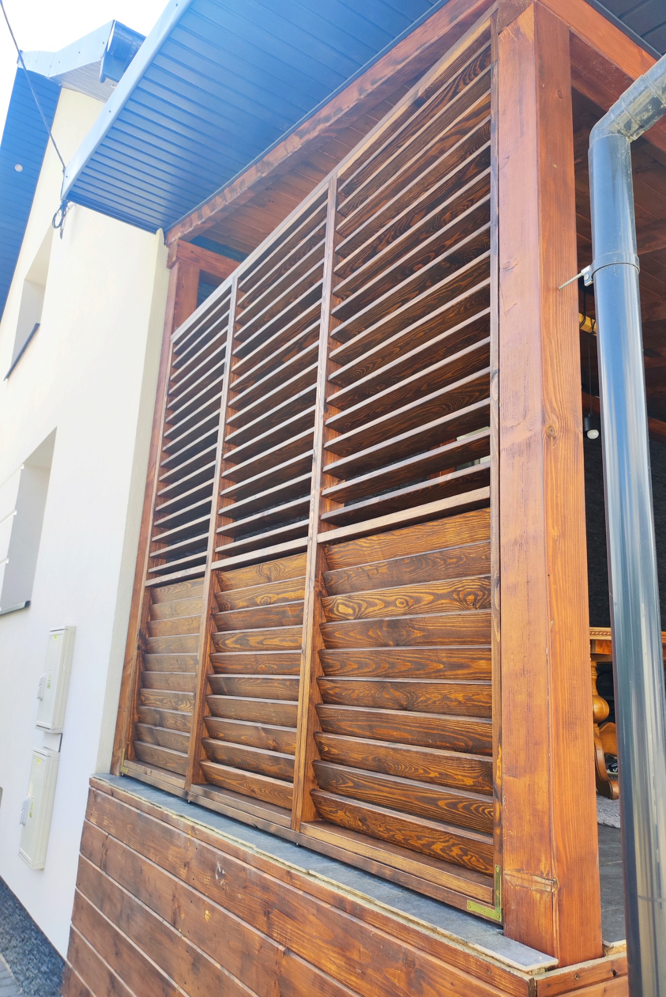Zabudowa drewniana tarasu z żaluzjami w kolorze ciemnego brązu, widok z zewnątrz, z elementami elewacji budynku w tle.