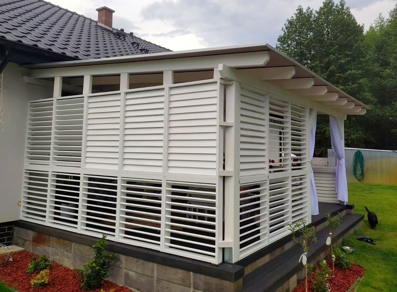 Nowoczesna, biała konstrukcja z drewnianymi żaluzjami, przylegająca do domu z ciemnym dachem, z widocznym fragmentem ogrodu i białymi zasłonami.