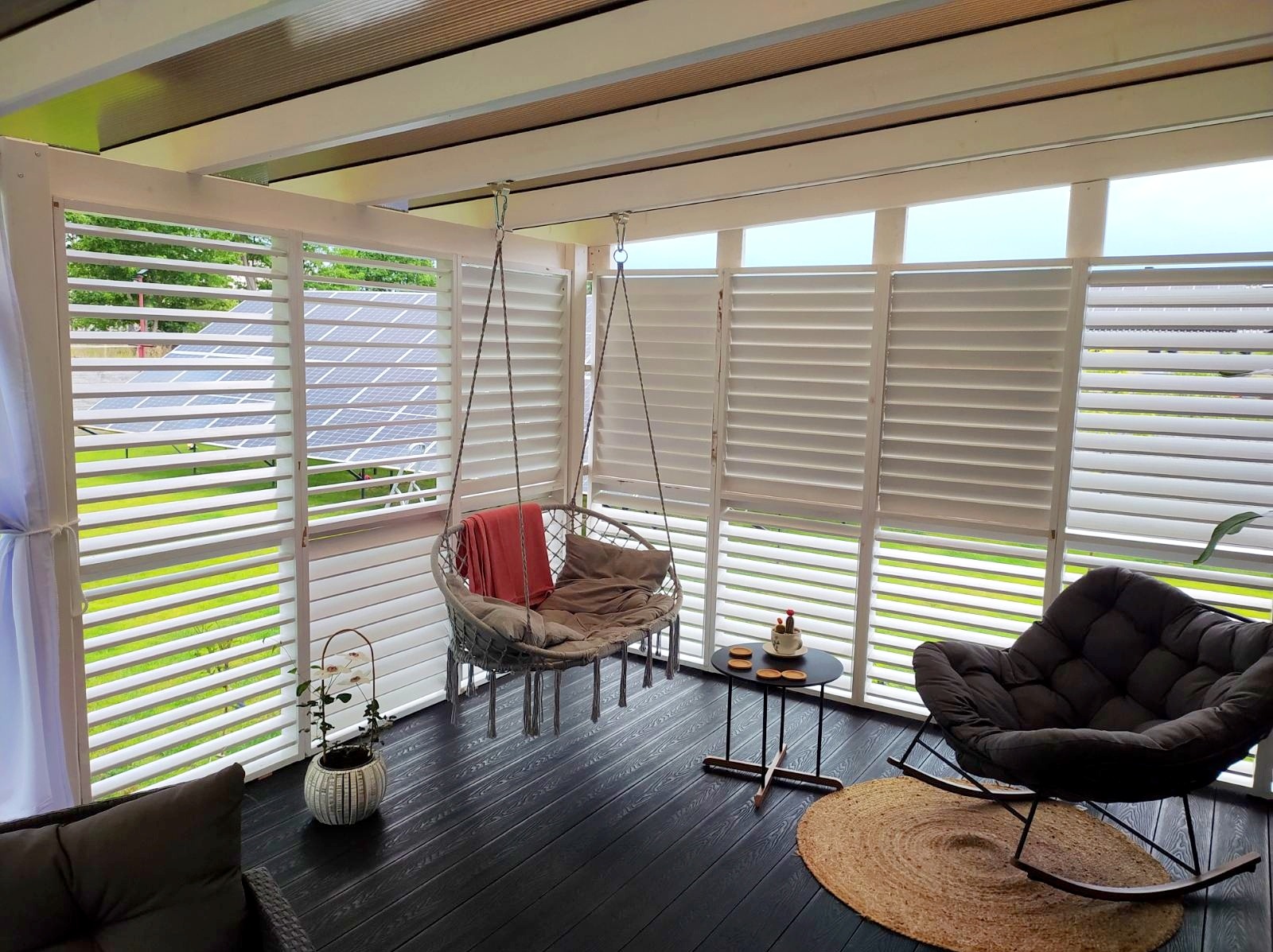 Zadaszona przestrzeń z drewnianą podłogą, wiszącym fotelem, roletami zewnętrznymi i widokiem na panele słoneczne.