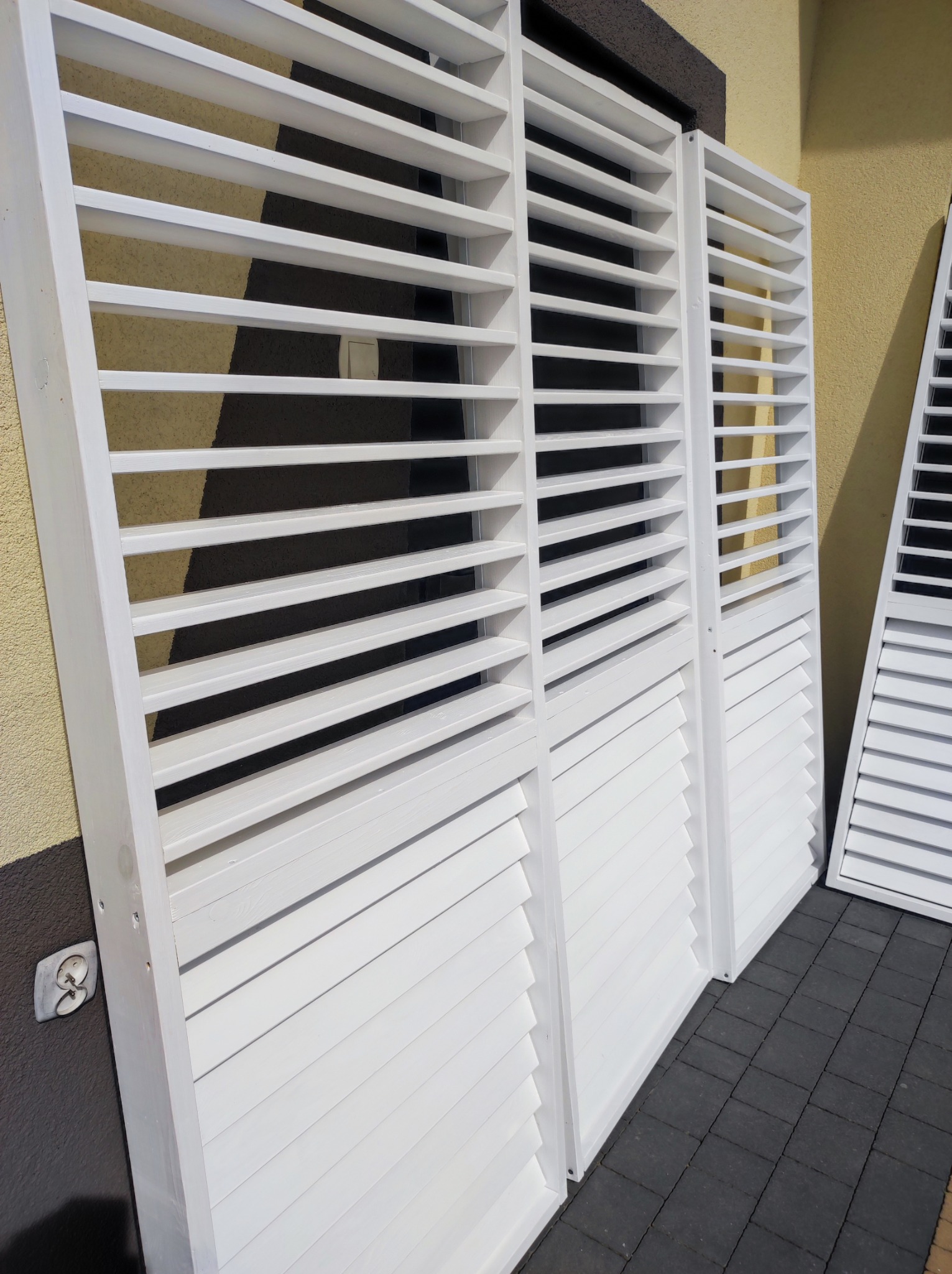 Białe, drewniane okiennice lamelowe, ustawione na kostce brukowej, gotowe do montażu, z widocznym gniazdkiem elektrycznym na ścianie budynku.