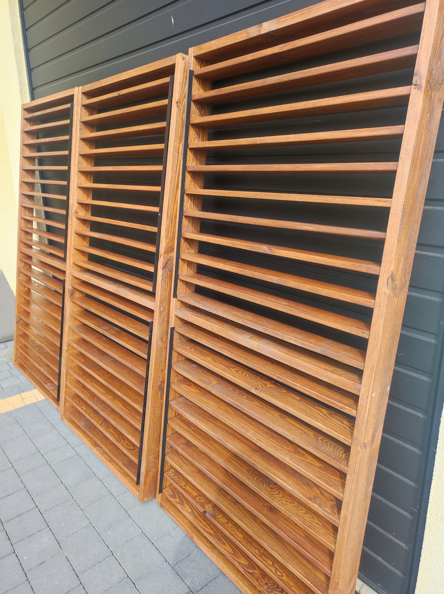 Trzy drewniane, brązowe konstrukcje z poziomymi lamelami, oparte o szarą fasadę z blachy trapezowej i ułożone na szarej kostce brukowej.