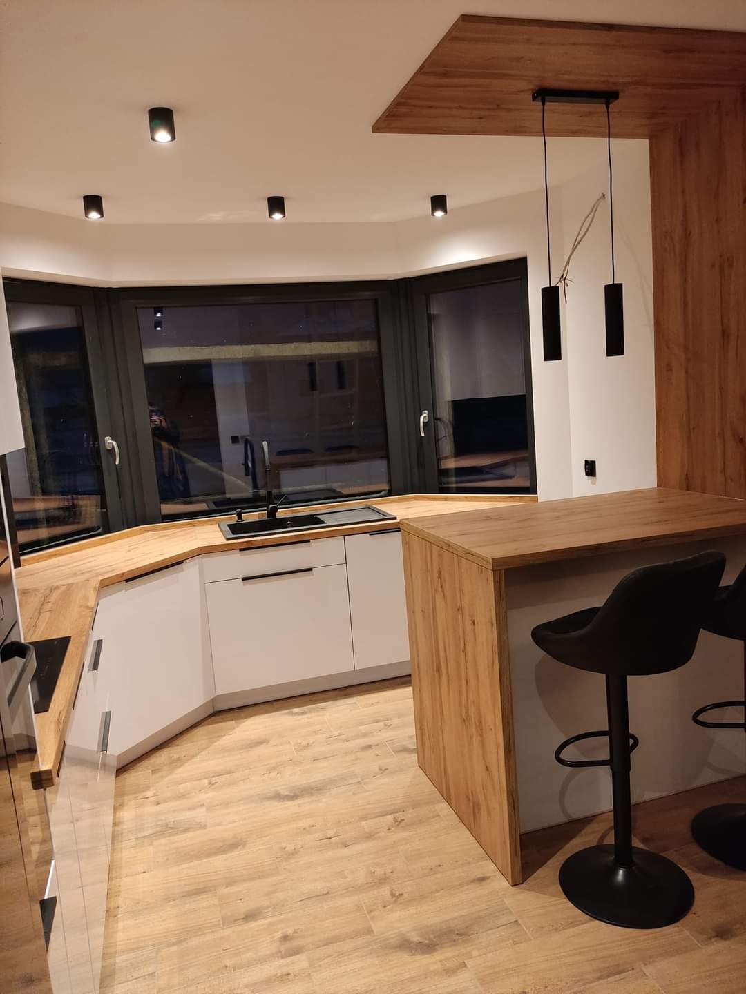 Nowoczesna kuchnia z białymi szafkami, drewnianym blatem i barkiem, oświetlona wiszącymi lampami i punktowymi reflektorami, z widocznym oknem wykuszowym.