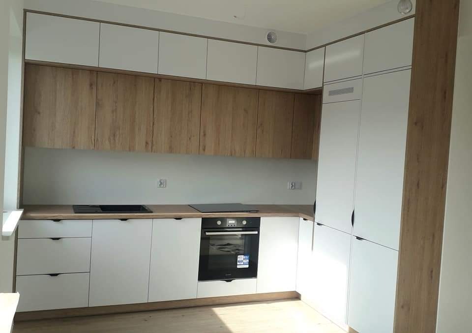 Nowoczesna kuchnia w kształcie litery L z białymi szafkami dolnymi i górnymi, z drewnianymi frontami szafek wiszących, czarnym piekarnikiem i płytą indukcyjną, oraz drewnianym blatem.