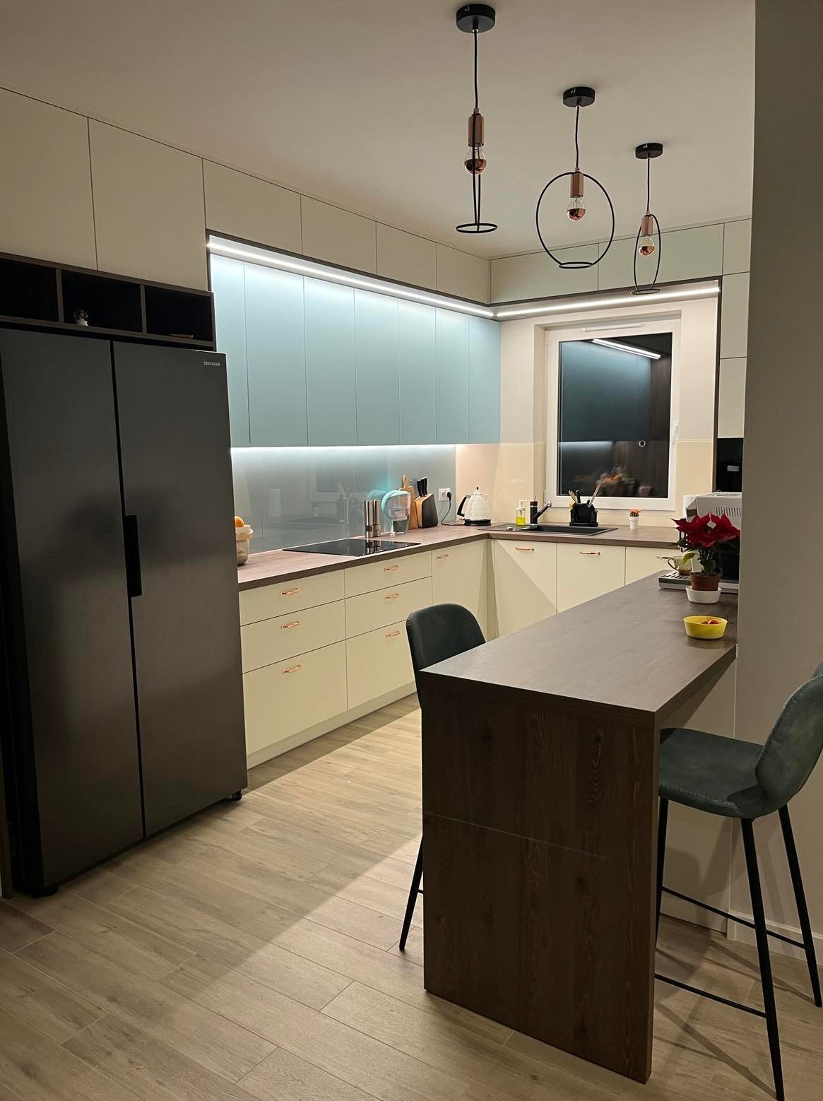 Nowoczesna kuchnia z jasnymi szafkami, turkusowym panelem ściennym, drewnianym blatem i barkiem oraz trzema designerskimi lampami wiszącymi.