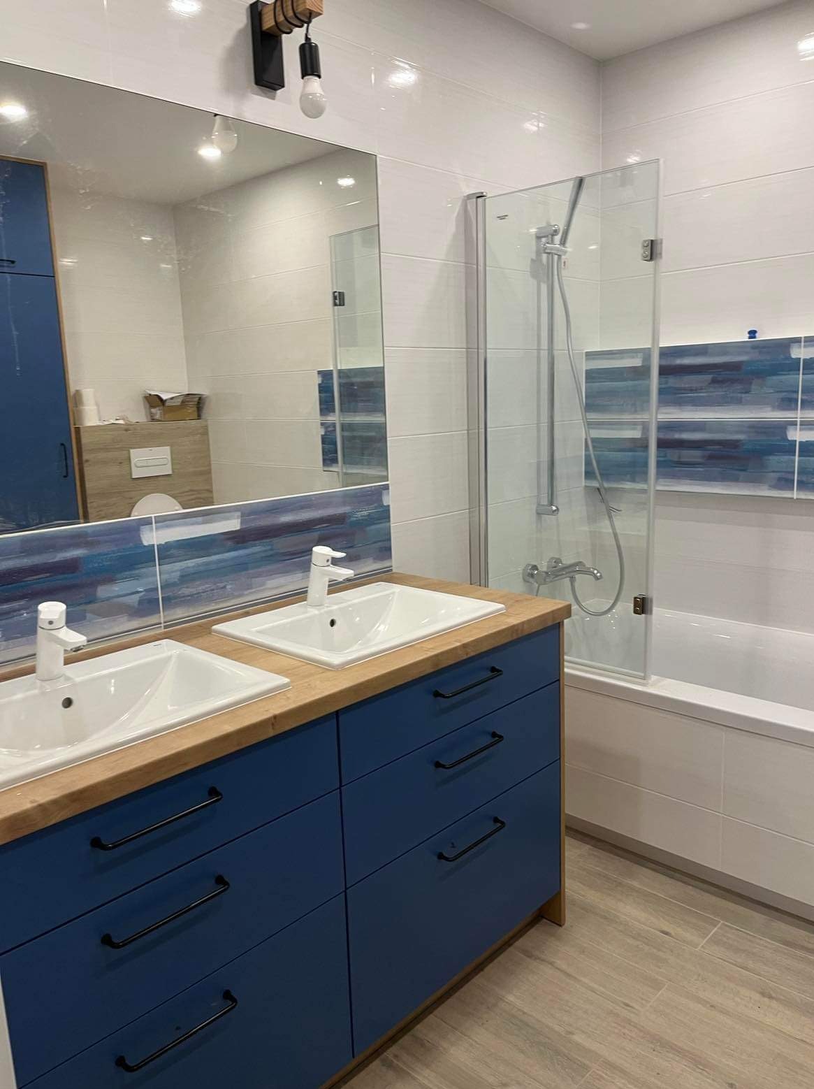 Nowoczesna łazienka z podwójną umywalką na drewnianym blacie i niebieskimi szafkami, kabina prysznicowa ze szklanymi drzwiami, białe płytki na ścianach z dekoracyjnym pasem niebieskich płytek.