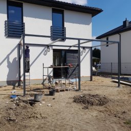 Spaw Kam - Montaż metalowej konstrukcji z oświetleniem przy białym budynku z balkonami, pracownik na drabinie, teren przygotowany pod budowę.