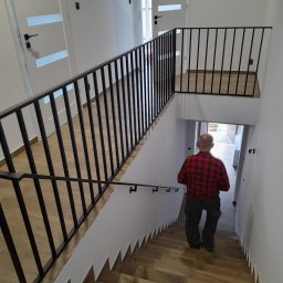 Spaw Kam - Wnętrze domu z drewnopodobnymi schodami i czarną metalową balustradą. Mężczyzna w czerwonej koszuli w kratę schodzi po schodach w dół.