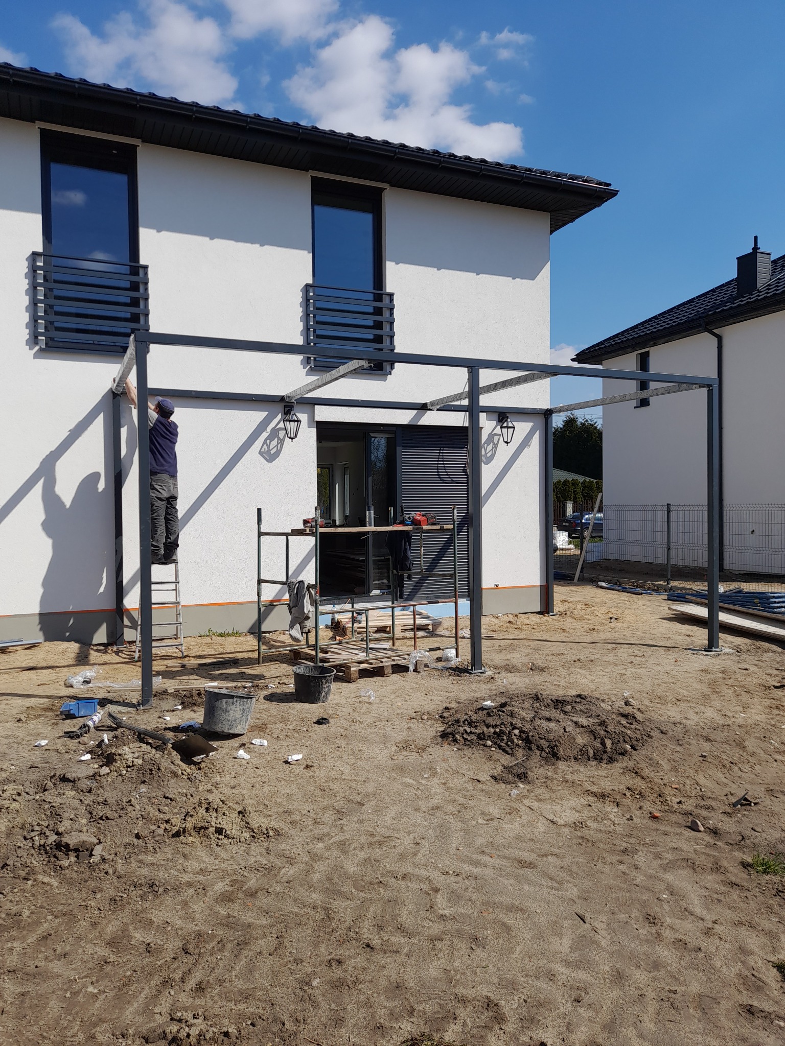 Montaż metalowej konstrukcji z oświetleniem przy białym budynku z balkonami, pracownik na drabinie, teren przygotowany pod budowę.