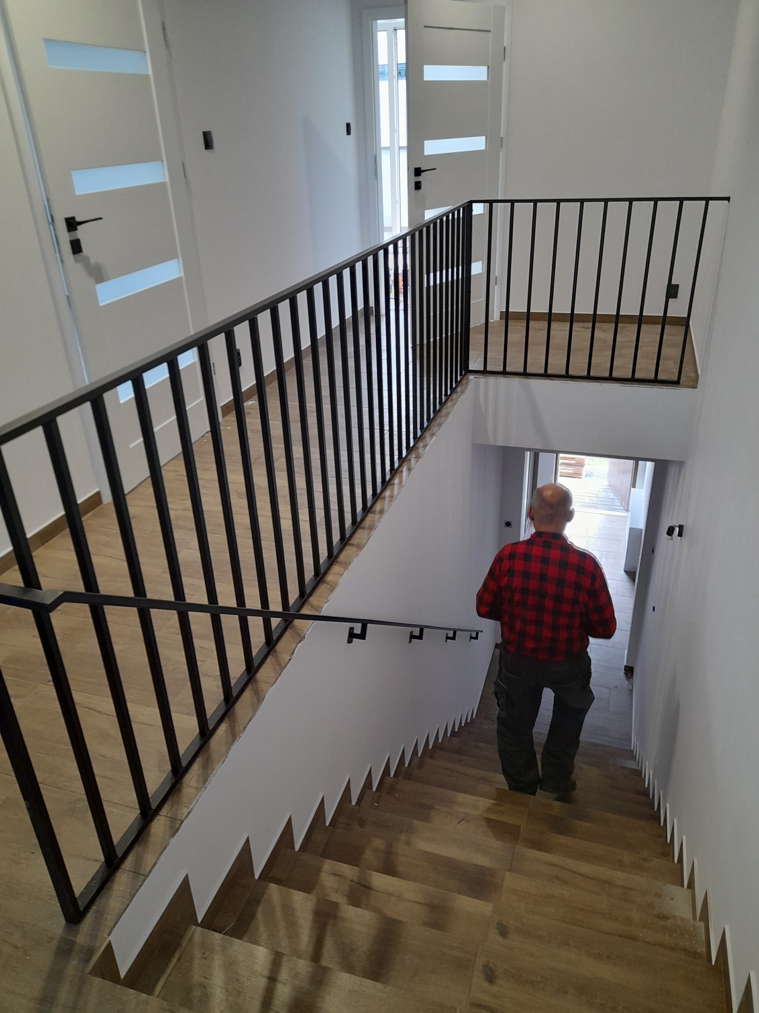 Wnętrze domu z drewnopodobnymi schodami i czarną metalową balustradą. Mężczyzna w czerwonej koszuli w kratę schodzi po schodach w dół.