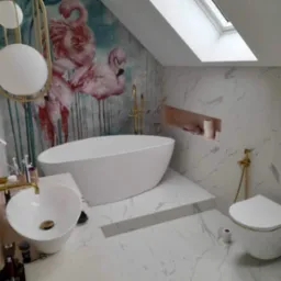 Elegancka łazienka na poddaszu z marmurowymi płytkami, wolnostojącą wanną, umywalką nablatową z złotą armaturą, toaletą ze złotymi akcentami i fototapetą z motywem flamingów.