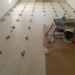 Ułożone płytki podłogowe z systemem poziomowania, widoczna drabina, pojemnik z zaprawą i butelka wody w trakcie prac glazurniczych.