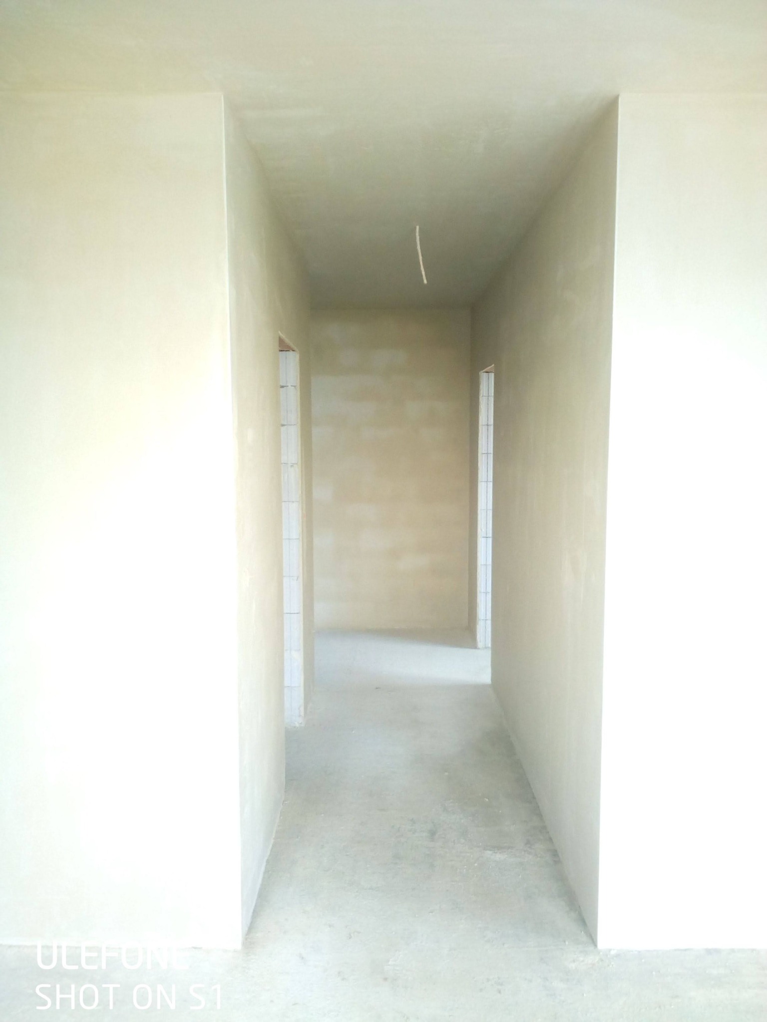 Korytarz w surowym stanie po nałożeniu jasnej, gładkiej warstwy tynku, widoczne przygotowane ościeżnice drzwiowe i wystający kabel z sufitu.