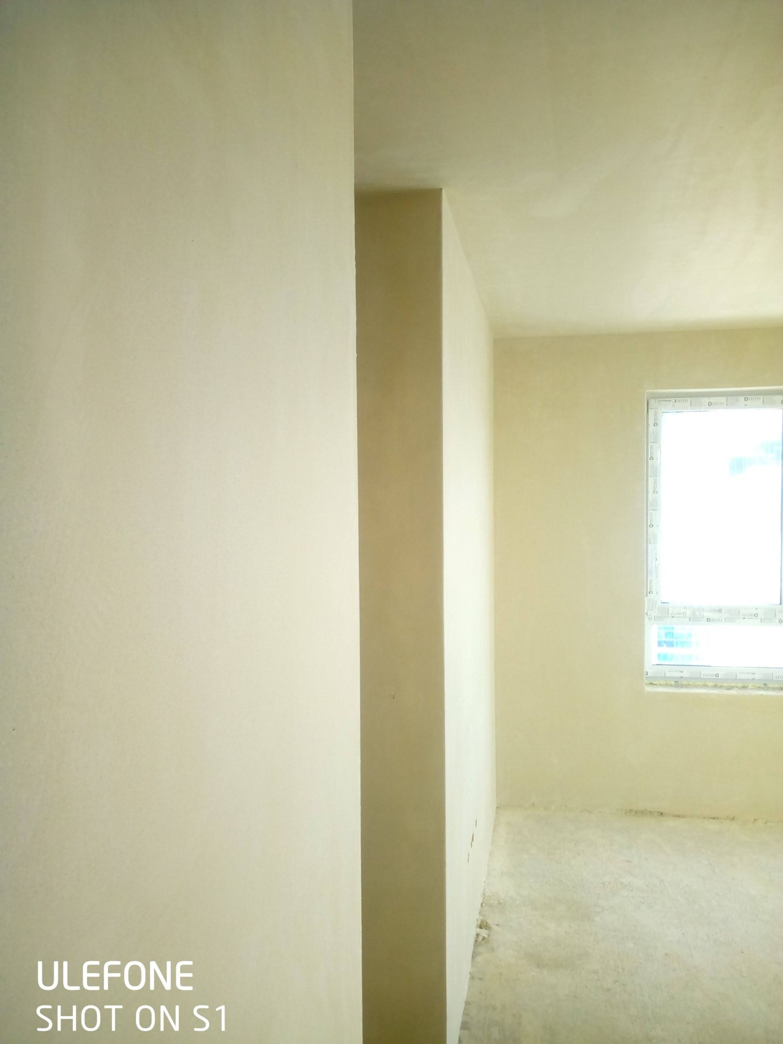 Świeżo otynkowane, gładkie ściany w jasnym odcieniu beżu, widoczne z perspektywy narożnika pomieszczenia z oknem.