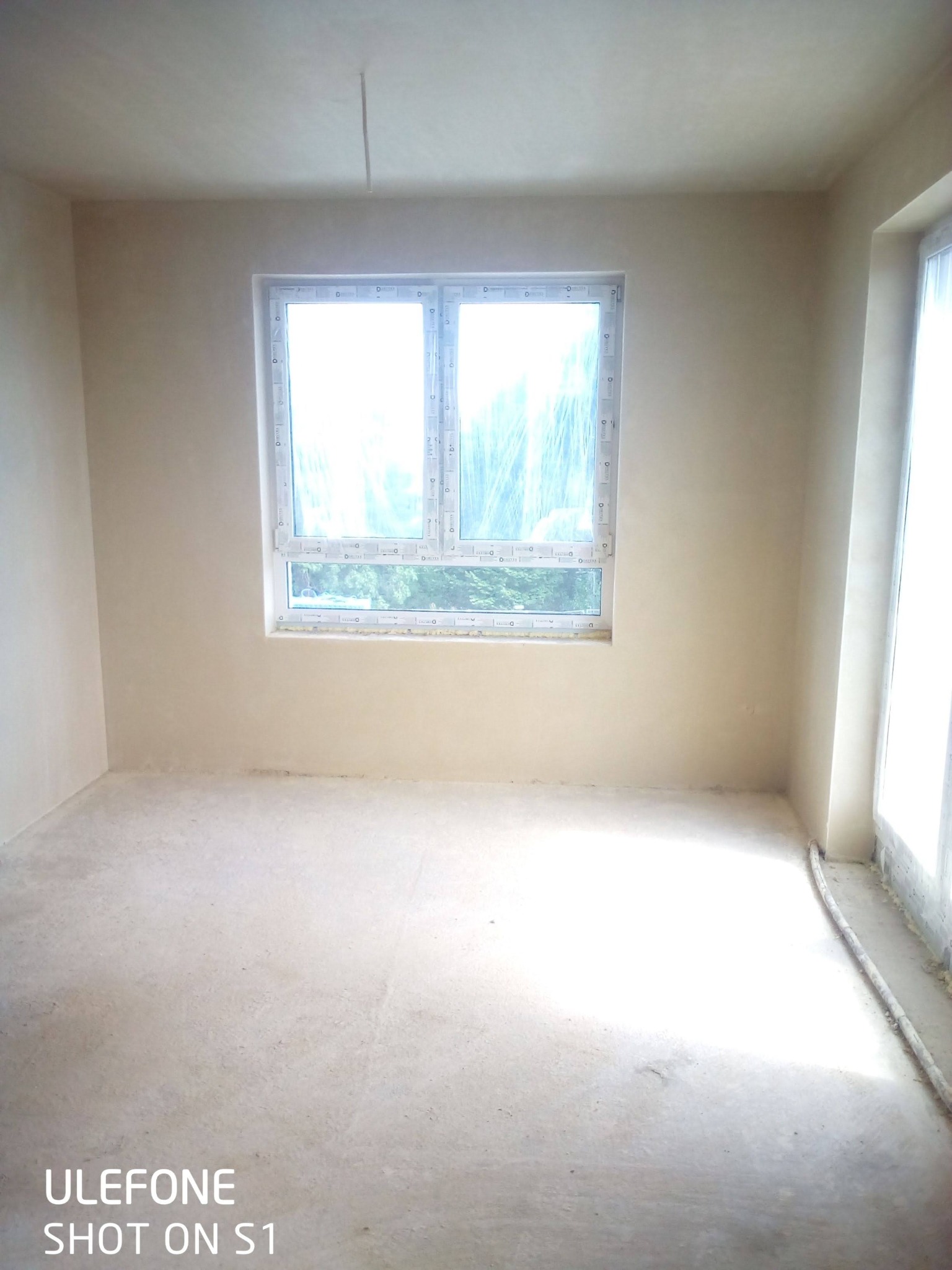 Pokój po nałożeniu gładzi, okna zabezpieczone folią, widoczne niedoskonałości na ścianach i podłodze, przygotowany do dalszych prac wykończeniowych.