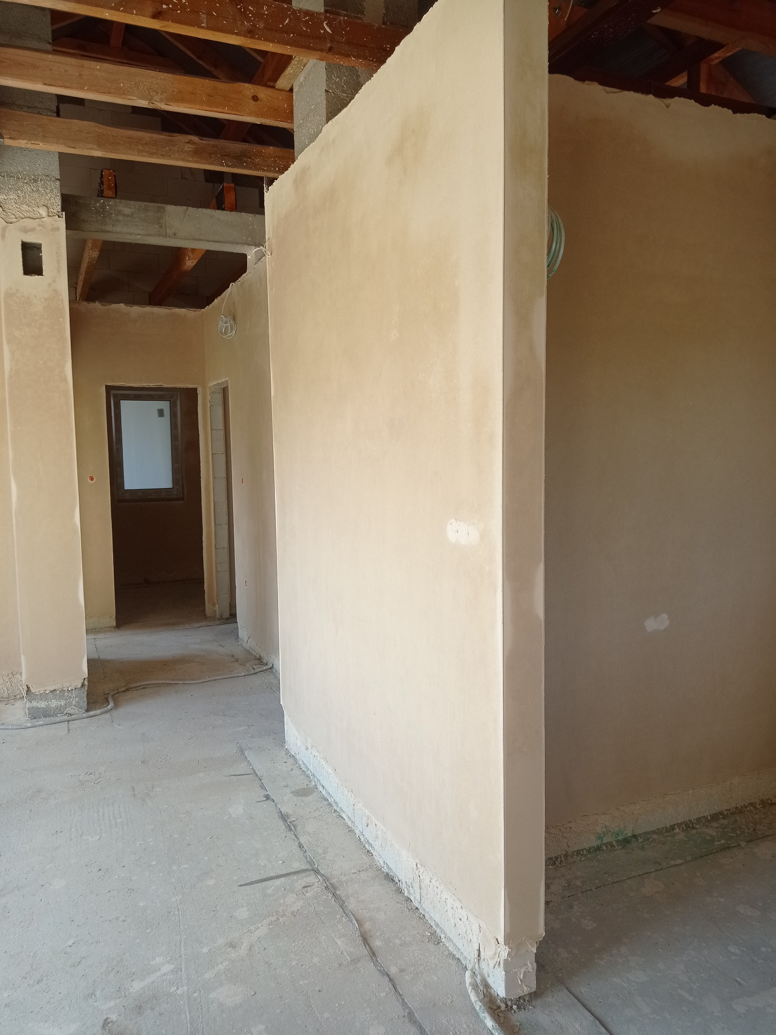 Świeżo nałożone tynki maszynowe na ścianach wewnętrznych budynku w trakcie budowy, widoczne drewniane elementy konstrukcji stropu i okno w głębi.